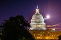 Full moon and clouds stand in sky behind marble dome of United States capitol building at night with trees silhouetted in national