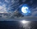 Full moon on cloudy fluffy dark night starry sky at sea and moonlight reflection on water on horizon city silhouette nature landsc
