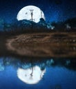 Full moon with milky way stars hidden behind the mountain and trees at the night time on sea with lunar reflection on the water.