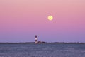 Full moon rising through the Belt of Venus behind a tall stone lighthouse at sunset.