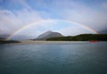 Waterfront with full rainbow on shore framing mountain in sunny blue sky and kayaks in water