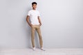 Full size photo of brunet optimistic curly guy stand wear white t-shirt trousers sneakers isolated on grey color