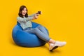 Full size photo of cheerful woman striped sit on armchair look empty space hold joystick play game isolated on yellow