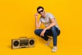 Full size photo of nice brunet young guy sit near boombox wear t-shirt glasses jeans sneakers on yellow