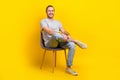 Full size photo pleasant friendly man dressed gray t-shirt denim pants clothes sitting on chair isolated on yellow color