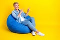 Full size photo of pretty young girl point look empty space sit beanbag dressed stylish denim outfit isolated on yellow Royalty Free Stock Photo