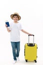 Teenage boy traveler in casual t-shirt, jeans and straw hat, walks with boarding pass and suitcase on white background. Royalty Free Stock Photo