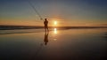 From full sun at sunset silhouetted fisherman with his very long rod on reflecting wet beach sand