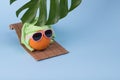 Fun idea made with an orange in sunglasses wearing a towel on his head lying on a sunbed and a monstera leaf on a blue background