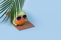 Fun idea made with an orange in sunglasses wearing a towel on his head lying on a sunbed and a palm leaf on a blue background