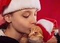 Fun kid girl playing holding and kissing with love the orange maine coon cat in christmas santa claus cap  on red background. Royalty Free Stock Photo