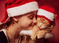 Fun kid girl playing the orange maine coon cat in christmas santa claus cap  on red background. Closeup portrait Royalty Free Stock Photo