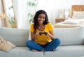 Fun stay home activities. Smiling black woman with joystick playing online video games on couch, full length portrait