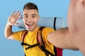 Funky millennial guy with camping gears taking selfie and waving to smartphone camera over blue studio background