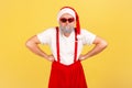 Funny adult man with grey beard in sunglasses, santa claus hat and pants with suspenders standing with arms akimbo Royalty Free Stock Photo