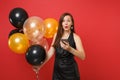 Funny amazed young woman in little black dress celebrating, holding air balloons and using mobile phone isolated on red Royalty Free Stock Photo