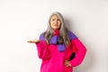 Funny asian mother with grey hair complaining, shrugging and looking left confused, pointing hand at something strange Royalty Free Stock Photo