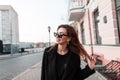Funny attractive happy young hipster woman with a positive smile in a fashionable black coat in stylish dark sunglasses stand