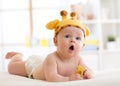 Funny baby boy in giraffe hat lying on his belly in nursery. Little kid lies on bed with opened mouth and holds soother