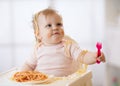 Funny baby eating noodle. Grimy kid girl eats spaghetti with fork sitting on table at home