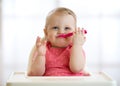 Funny baby with spoon in her mouth. Beautiful child girl sitting in high chair and waiting food. Nutrition for kids Royalty Free Stock Photo