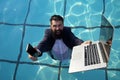 Funny bearded business man in suit using phone and laptop in swimming pool. Concept of young people working mobile Royalty Free Stock Photo