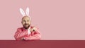 Funny bearded man in Easter Bunny ears pointing at pink advertising copy space background