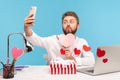 Funny bearded man office worker making selfie pressing lips in kiss and holding paper pink heart, sitting at workplace all covered Royalty Free Stock Photo