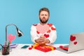 Funny bearded man sitting at workplace all covered with sticky hearts and sending air kisses holding pink heart greeting card, Royalty Free Stock Photo