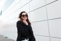 Funny beautiful young hipster woman in a black stylish coat in vintage white jeans in dark trendy sunglasses goes and laughs Royalty Free Stock Photo