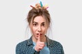 Funny beautiful young woman with wild hair and colorful markers in hair, holding her index finger to her lips to keep silence, Royalty Free Stock Photo