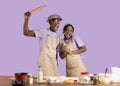 Funny black guy with girlfriend wearing aprons, holding kitchen utensils, having fun while cooking on violet background Royalty Free Stock Photo