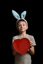 Funny blond boy in hare ears holds heart shape red box on black background. Love and family concept. Vertical frame