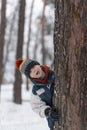 Funny boy peeks out from behind tree. Child in knitted hat walks through winter snow-kept park Royalty Free Stock Photo