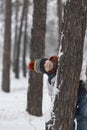 Funny child boy hides behind tree in winter forest. Boy is dressed in warm hat and winter jumpsuit. Vertical frame