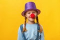 Funny child clown, hat and a red nose. Cheerful little girl on a yellow background. April 1. April Fools Day. Copy space