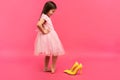 Funny child girl fashionista in dress going to put on big mother`s yellow shoes on colored background. Royalty Free Stock Photo