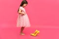 Funny child girl fashionista in dress going to put on big mother`s yellow shoes on colored background. Royalty Free Stock Photo