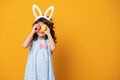 Funny child girl wearing bunny ears and holding up a colorful Easter egg in front of her eye on yellow background Royalty Free Stock Photo