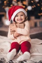 Funny child girl 3-4 year old holding Christmas present box wearing santa claus hat and suit sitting over tree with glowing lights Royalty Free Stock Photo