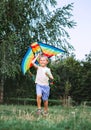 Funny childhood with lovely toys concept image. Cheerful little blonde hair boy running with a multicolored kite on the city park Royalty Free Stock Photo