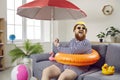 Funny chubby man having fun sitting on sofa in living room with inflatable swimming circle.