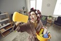 Funny crazy young housewife in curlers and skin care face mask tidying up her house