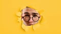 Funny curious smart caucasian young guy squints, takes off his glasses and looks into hole in yellow paper