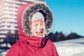 Funny cute Caucasian smiling girl in red jacket parka squinting eyes from bright sun. Happy kid with snow on face at cold winter