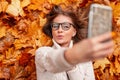 Funny cute happy young woman hipster in stylish glasses in a knitted sweater photographs herself on a mobile phone. Modern pretty Royalty Free Stock Photo