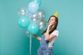 Funny cute young woman in birthday hat looking aside blowing in pipe, celebrating, holding colorful air balloons Royalty Free Stock Photo