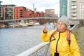 funny elderly woman in yellow jacket and hat points to Gutleitviertel district in Frankfurt, buildings on river Main shore, boats