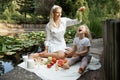 Funny family sitting on plaid with fruits, jug of milk on edge of pond. Young woman holding grapes over little girl. Royalty Free Stock Photo