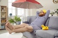 Funny fat man spends summer vacation at home sitting on sofa under fan and beach umbrella.